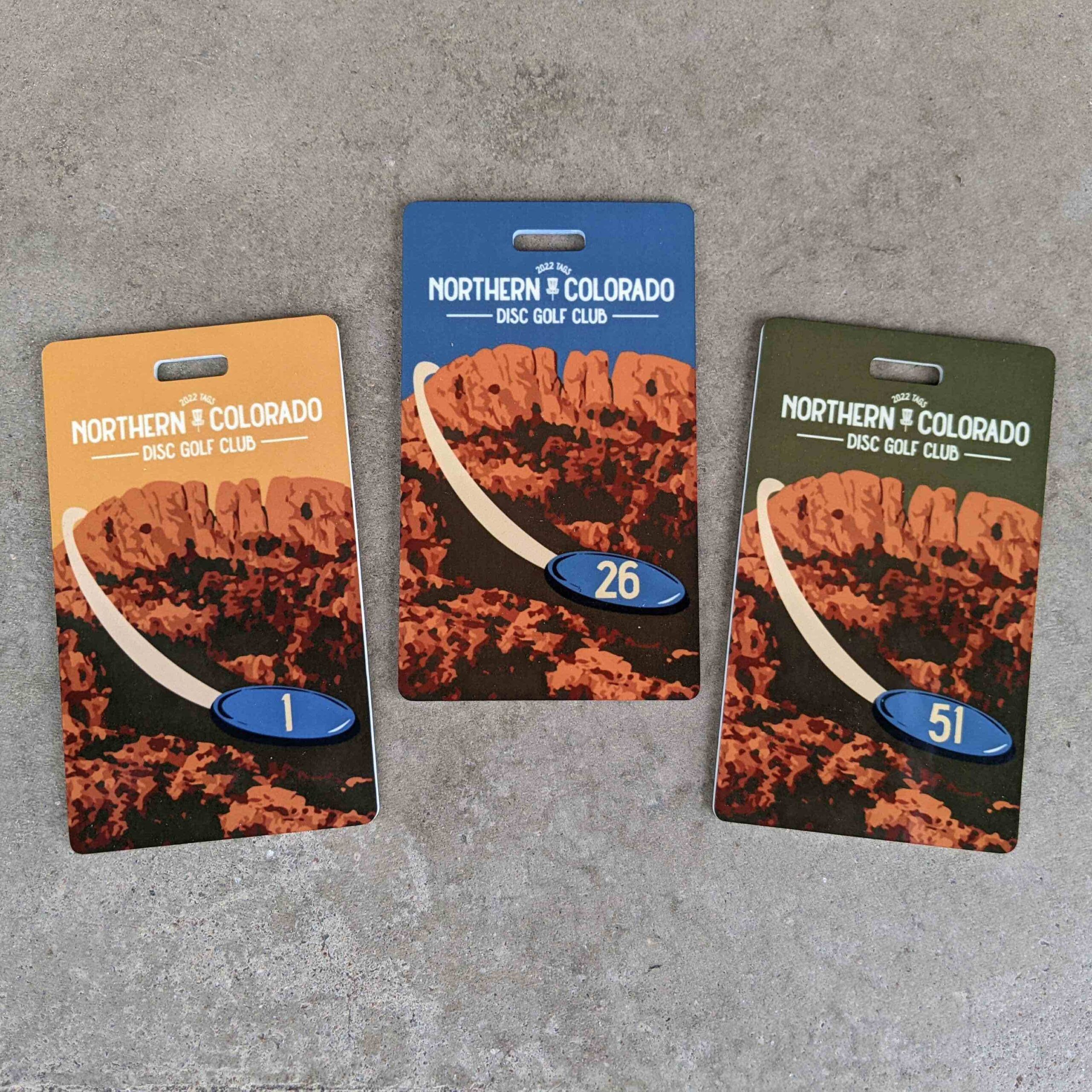 Image shows three rectangular plastic tags laying on pavement. Each shows Horsetooth rock with a blue disc flying into the foreground which has the tag number on it. The top of each tag says, "Northern Colorado Disc Golf Club 2022 Tags".
